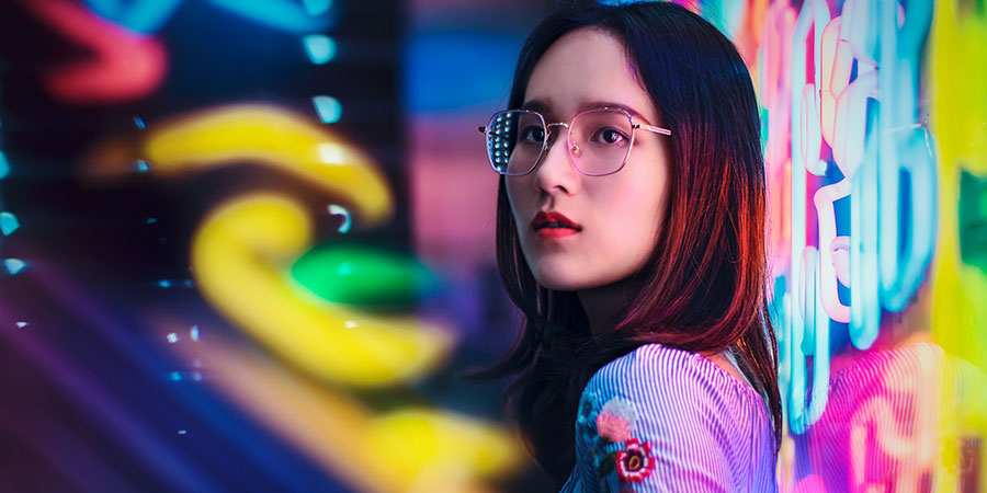 Asian woman wearing glasses standing near a neon sign